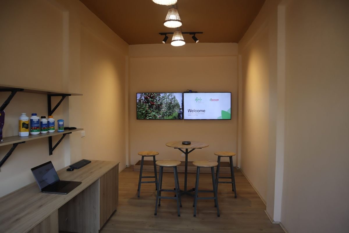 Inside a Qulgah centre: screens showing an orchard and a welcome slide, a table and stools, and a shelf of trial products.